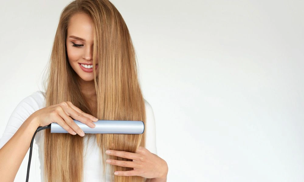 A smiling woman with long, straight blonde hair uses a silver flat iron to smooth a section, against a clean white background. The scene feels calm and polished.