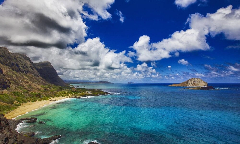 Turquoise water laps a sandy Hawaiian beach beneath green mountains and dramatic clouds-Best Places to Snorkel