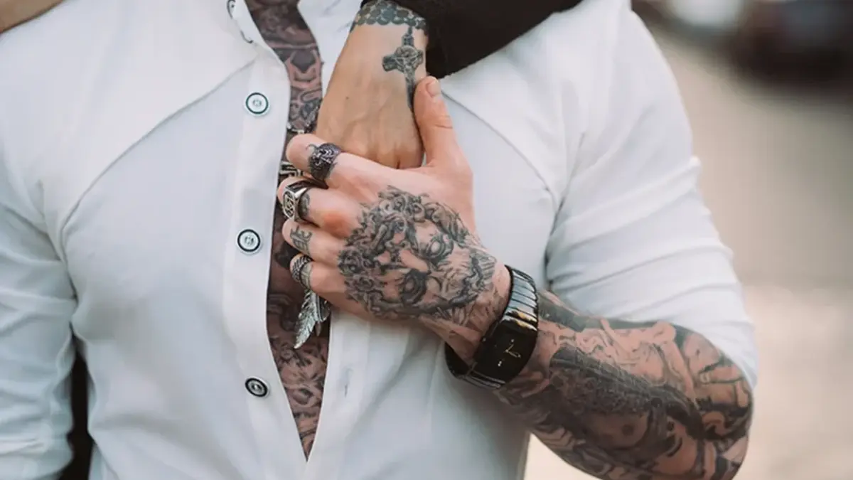 Close-up of a person in an open white shirt clasping tattooed hands at their chest, wearing rings and a dark watch; bold, edgy style.