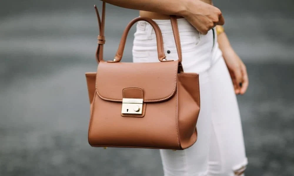 A woman in white jeans carries a structured tan leather handbag with a top handle and gold clasp, creating a polished, understated street-style look.