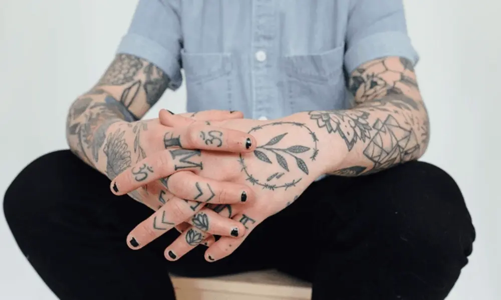Tattooed person in a light blue shirt and black trousers sits with hands clasped, displaying detailed floral and symbolic ink in a calm, minimalist portrait.