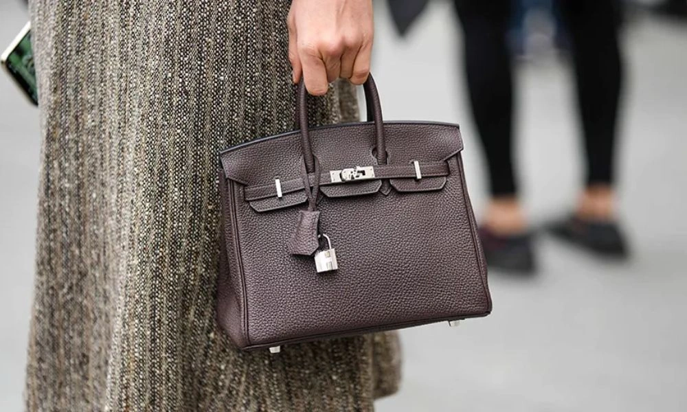 A person in a textured gray outfit carries a dark brown leather handbag with silver hardware and a small lock; blurred pedestrians suggest a busy city street.