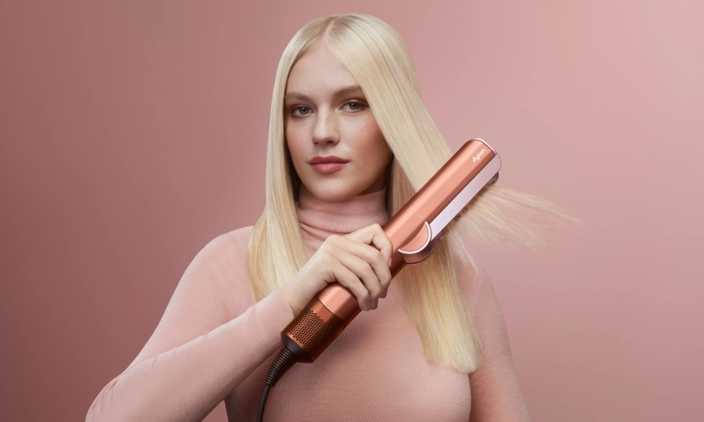 A blonde woman in a soft pink top holds a rose-gold hair straightener