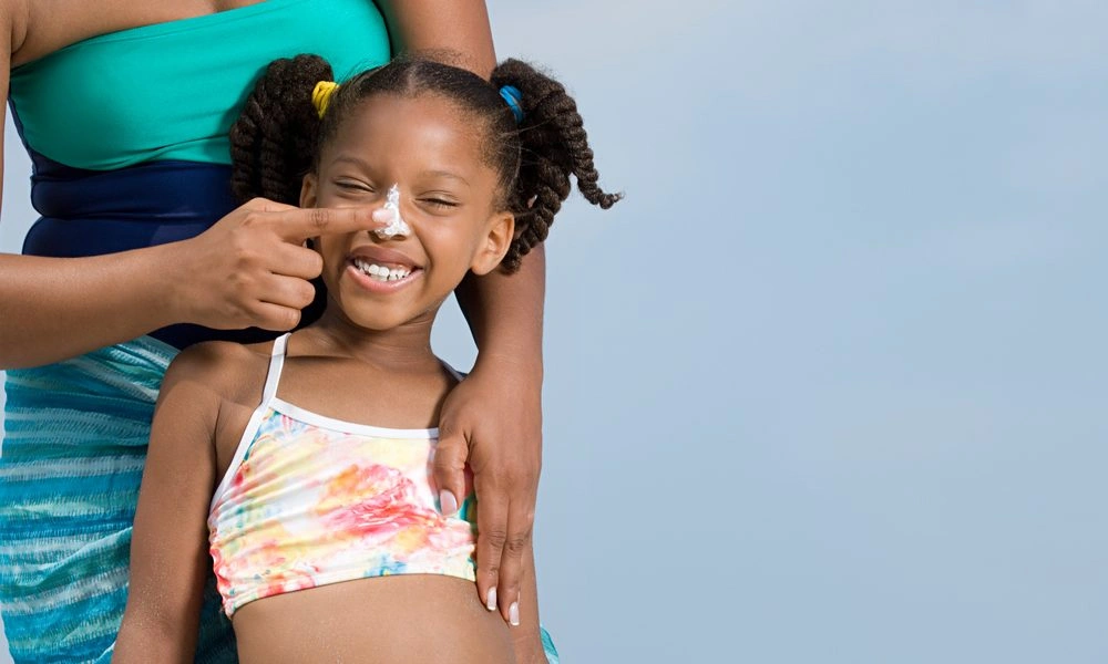 A smiling girl in a colorful swimsuit has sunscreen dabbed on her nose by an adult, sharing a playful, caring moment before sun exposure.