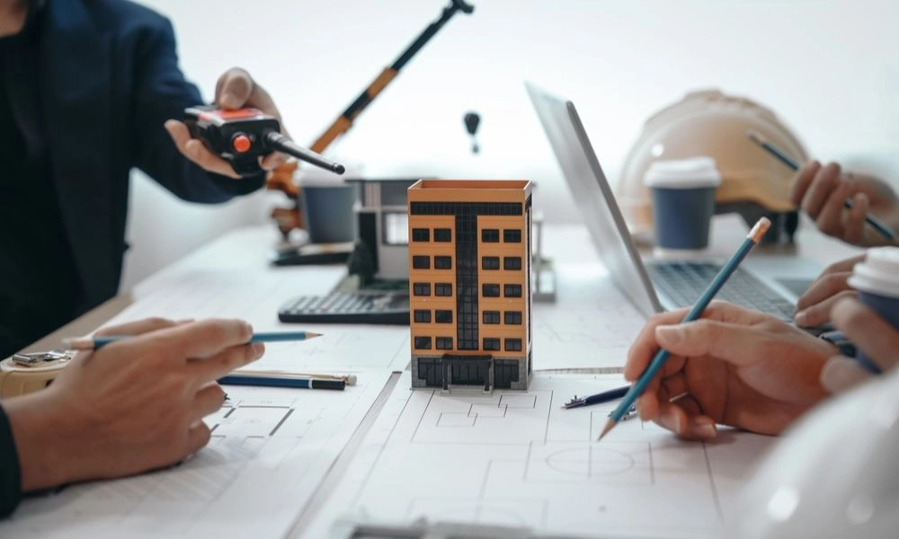 Architects gather around blueprints, pointing and sketching beside a small apartment building model, laptops and plans suggesting focused collaboration.