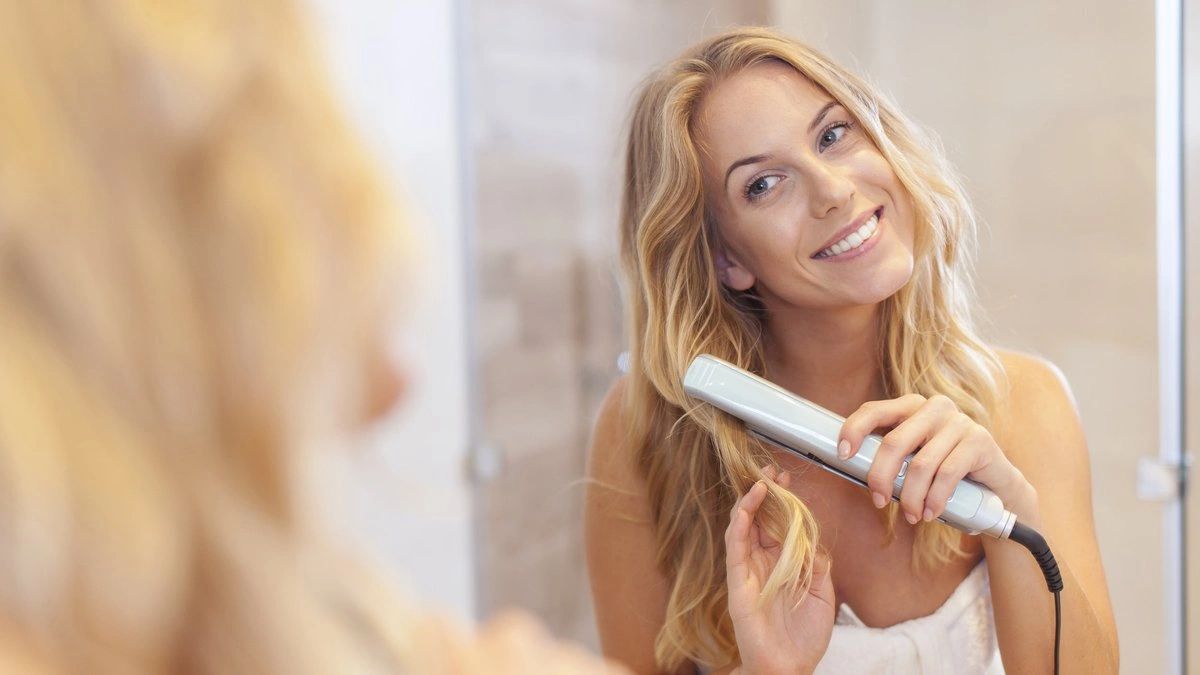 Smiling blonde woman straightens her hair with a flat iron in a bright bathroom, looking in the mirror; the scene feels relaxed and cheerful.