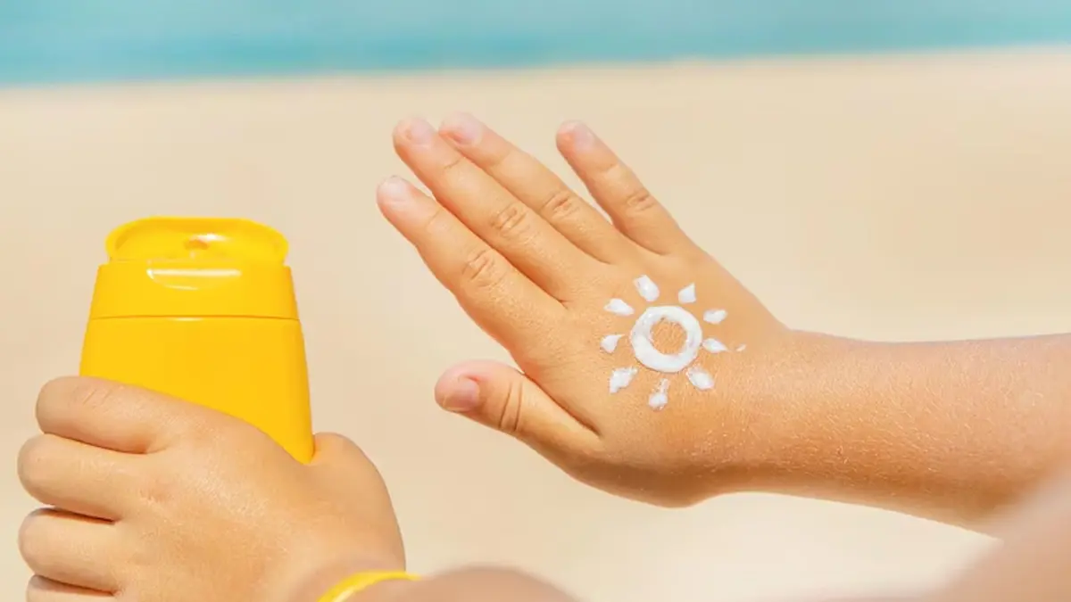 A child’s hand with white sunscreen sun and rays rests over a sandy beach beside a bright yellow sunscreen bottle, evoking sunny-day protection.