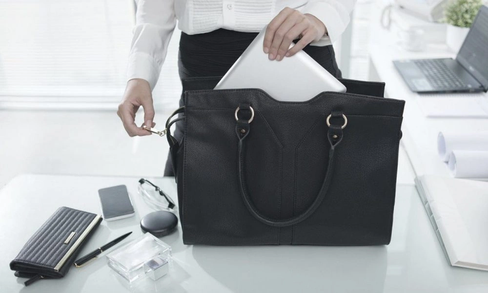 A businesswoman places a tablet in a structured black handbag on a bright office desk, surrounded by stationery and a laptop, conveying a polished, professional mood.