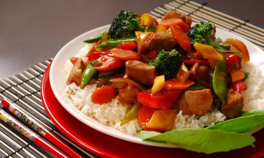 A colorful, appetizing beef and vegetable stir-fry with broccoli, peppers, and snap peas over white rice on a red-rimmed plate, with chopsticks nearby.