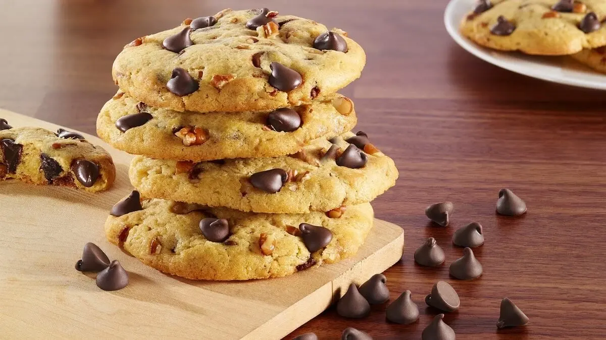 A stack of golden chocolate chip and nut cookies sits on a wooden board, with scattered chocolate pieces and more cookies behind, creating a warm, inviting feel.