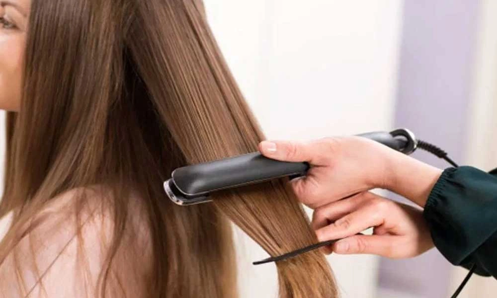A stylist uses a black flat iron and comb to straighten a section of a woman’s long brown hair in a salon, creating a calm, polished look.