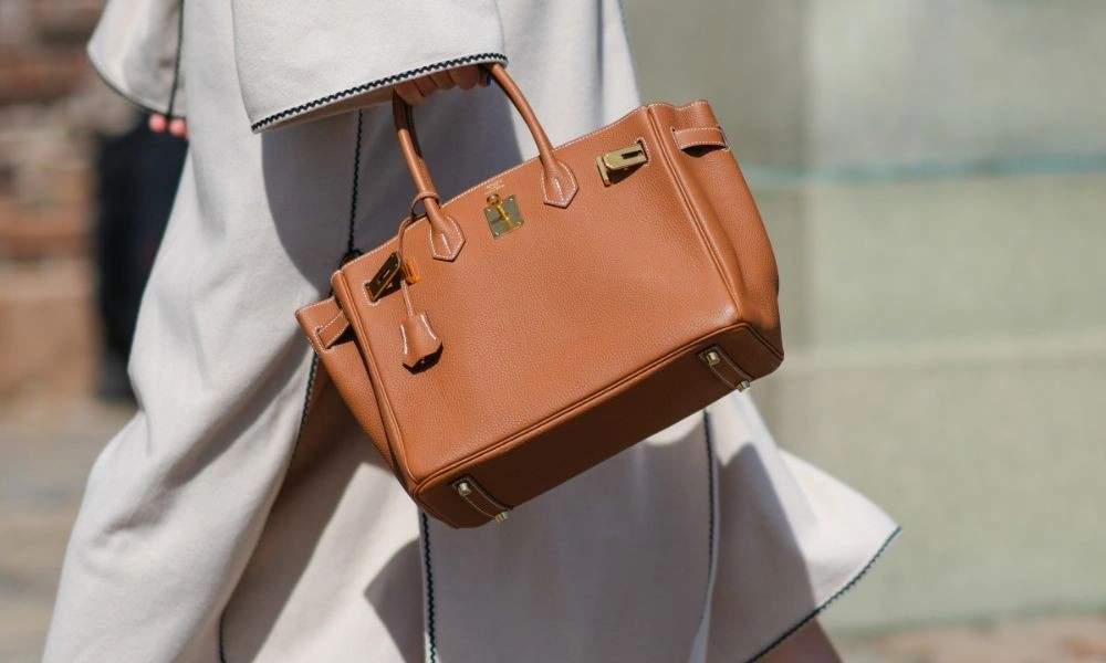 A woman in a flowing white outfit carries a structured cognac leather handbag with gold hardware outdoors, creating a polished, elegant fashion look.