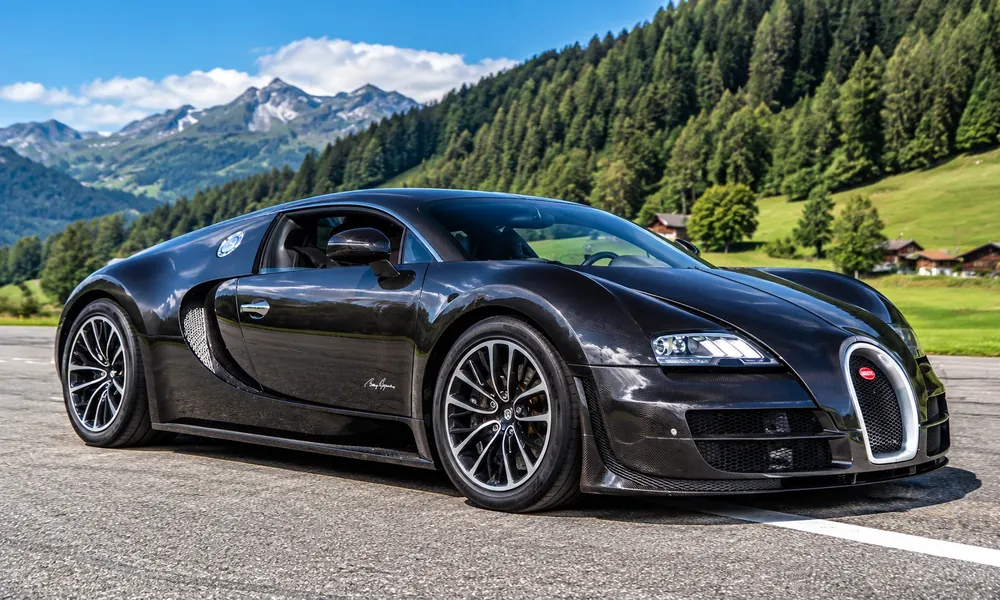 Black carbon fiber Bugatti Veyron Super Sport parked on a road with forested mountains behind it.