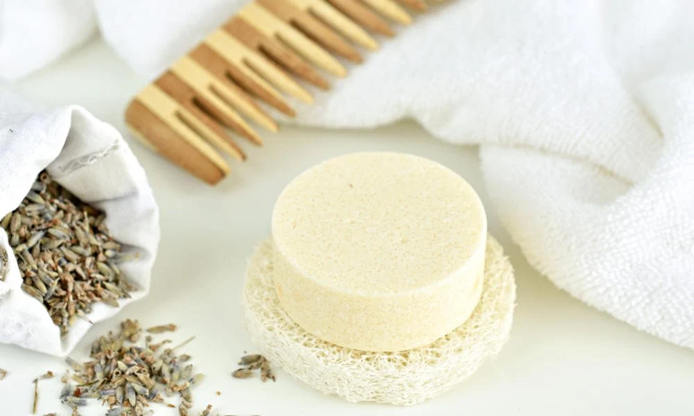 A pale round soap bar rests on a natural loofah beside a wooden bath brush, white towels, and dried lavender, creating a calm, spa-like mood.