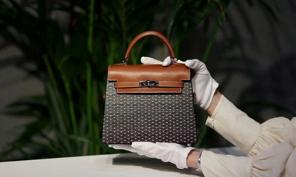 White-gloved hands display a structured brown leather handbag with a dark patterned body against leafy greenery, conveying a refined luxury-showroom feel.
