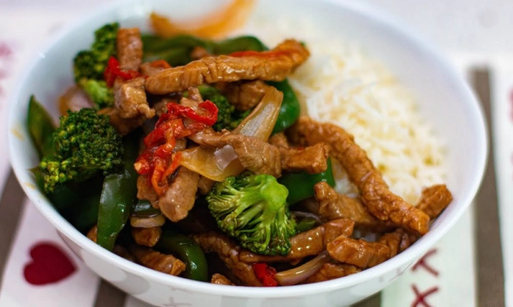 A white bowl of savory beef and broccoli stir-fry with red peppers and onions, served beside fluffy rice; a colorful, hearty meal-best dumpling recipes