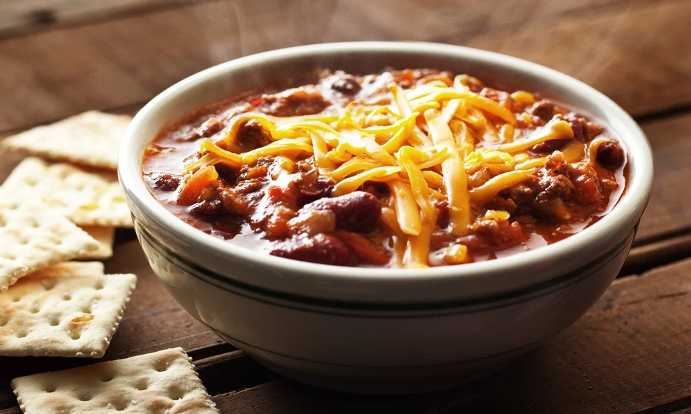 A bowl of hearty chili topped with shredded cheddar cheese sits on a rustic wooden table, with crisp crackers alongside for a cozy, appetizing meal.