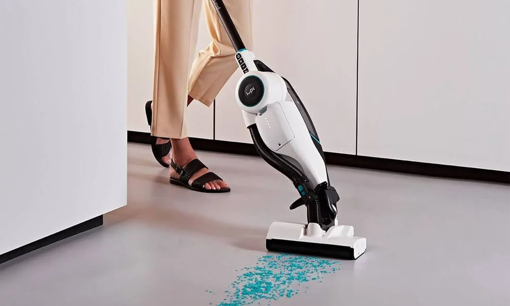 A person in tan trousers and sandals uses a white-and-black stick vacuum to clean turquoise debris from a gray floor in a bright, modern kitchen.