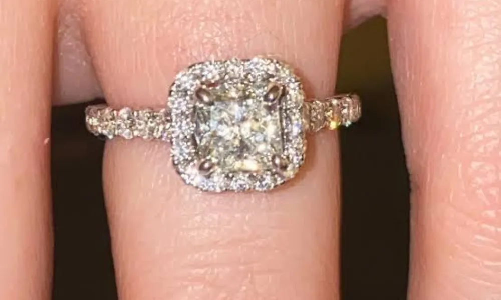 Luxury of Time by Diamonds International featured in a close-up photo of a cushion-cut diamond ring with a halo setting and pavé band worn on a finger. 