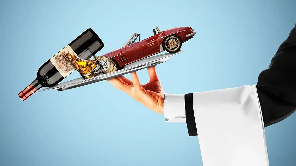 A server’s hand presents a tray with a wine bottle, filled glass, and red vintage sports car against a blue backdrop, creating a playful luxury mood.