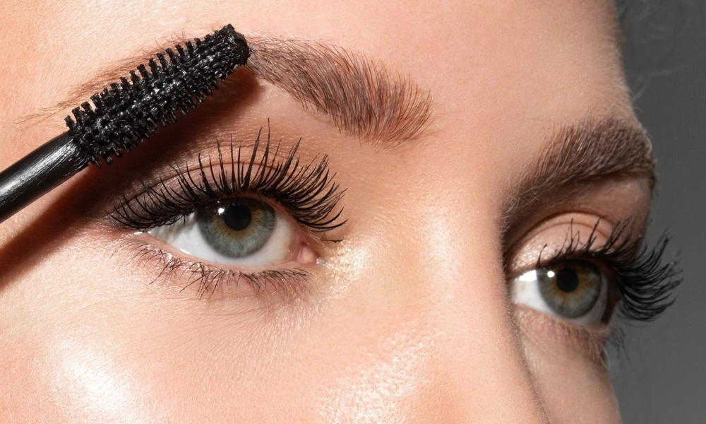 Close-up beauty image of a woman brushing her dark eyebrow with a mascara wand, showing long curled lashes and polished eye makeup in a bright studio.