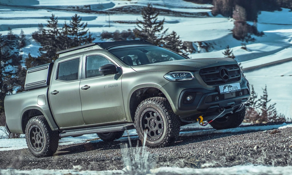 A rugged silver Mercedes-Benz pickup with off-road tires climbs a snowy, rocky trail, framed by evergreen trees and a wintry mountain landscape. Its stance feels adventurous.