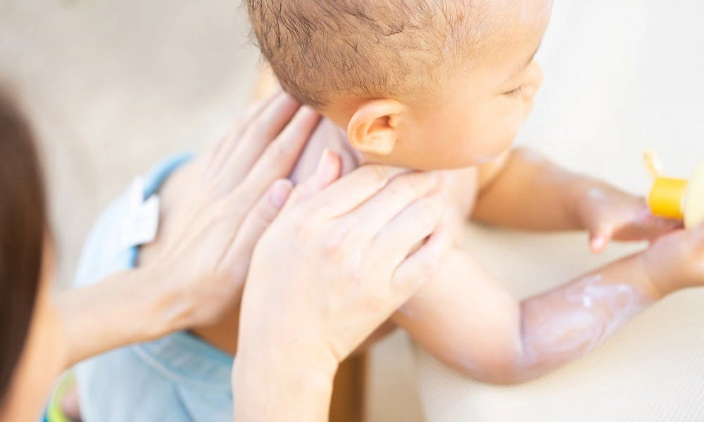 Caregiver gently supports a baby’s neck and chest during a bath as the infant reaches toward a yellow bath toy; a tender, reassuring moment.