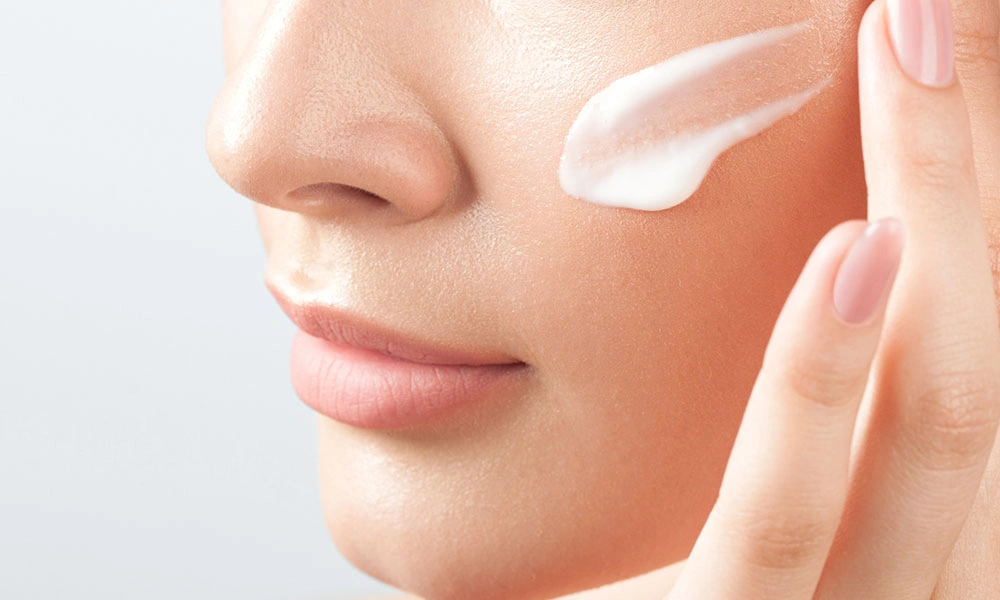 Close-up of a person applying a dab of white facial cream to their cheek, with fingertips resting on their skin; bright, clean lighting suggests gentle skincare.