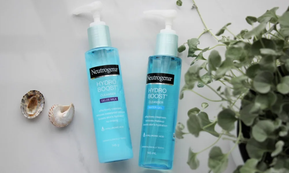 Two turquoise Neutrogena Hydro Boost cleanser pump bottles lie beside seashells and leafy greenery on a pale surface, creating a fresh, calming skincare scene.