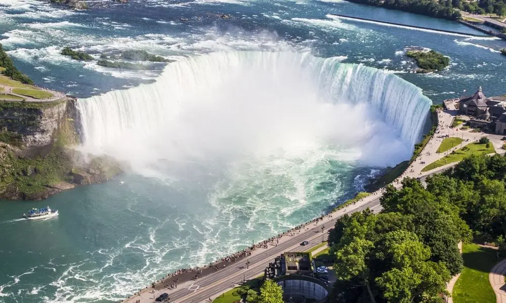 Aerial view of Niagara Falls, with water plunging over a broad curved cliff into misty turquoise rapids, beside a crowded walkway and green parkland.