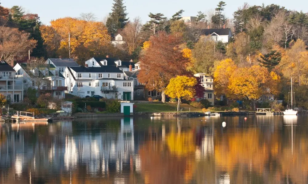 White lakeside homes and small docks sit beneath brilliant autumn trees, their gold and orange foliage reflected in the calm, still water-Prettiest Places in Canada