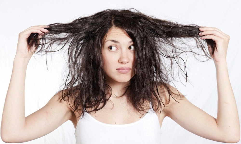 A woman in a white tank top stretches her long, tangled dark hair out to both sides, looking uncertain against a plain white background.