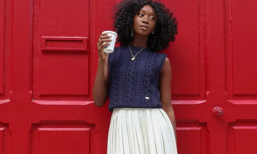 A young woman with natural curls holds a white cup before a vivid red paneled door, wearing a navy knit top and pleated white skirt; poised and calm-Football Game Outfit Ideas