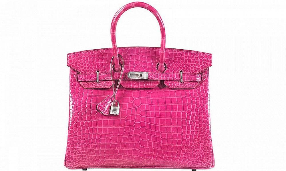 A bright fuchsia, crocodile-embossed leather handbag with rounded top handles, a silver clasp, and dangling lock charms, isolated on white.