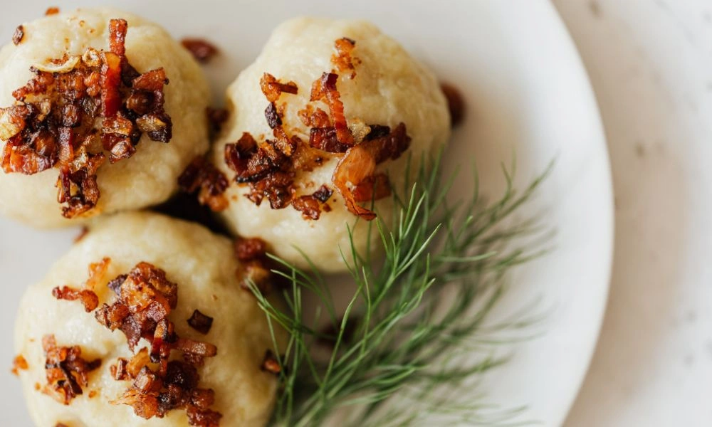 Three soft, golden dumplings topped with crispy fried onions and browned bits sit on a white plate beside fresh dill, creating a warm, appetizing dish.