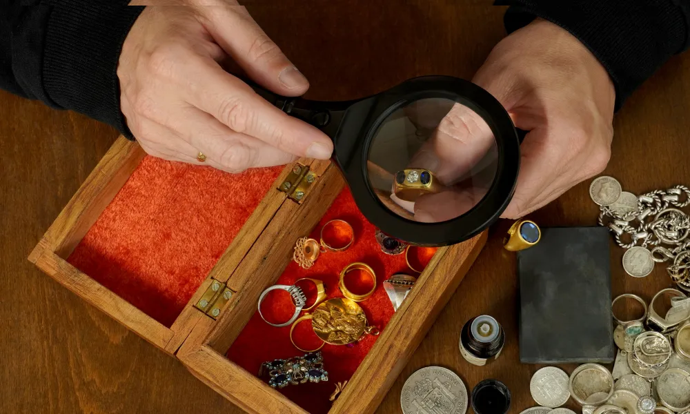 Hands use a magnifying glass to inspect a gold ring over a red velvet-lined wooden jewelry box, surrounded by rings, coins, and silver jewelry.