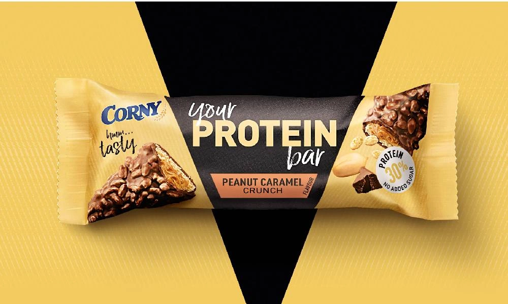 A Corny Protein peanut caramel crunch bar in a yellow wrapper, centered on a bright yellow background with a bold black triangle; energetic ad style.