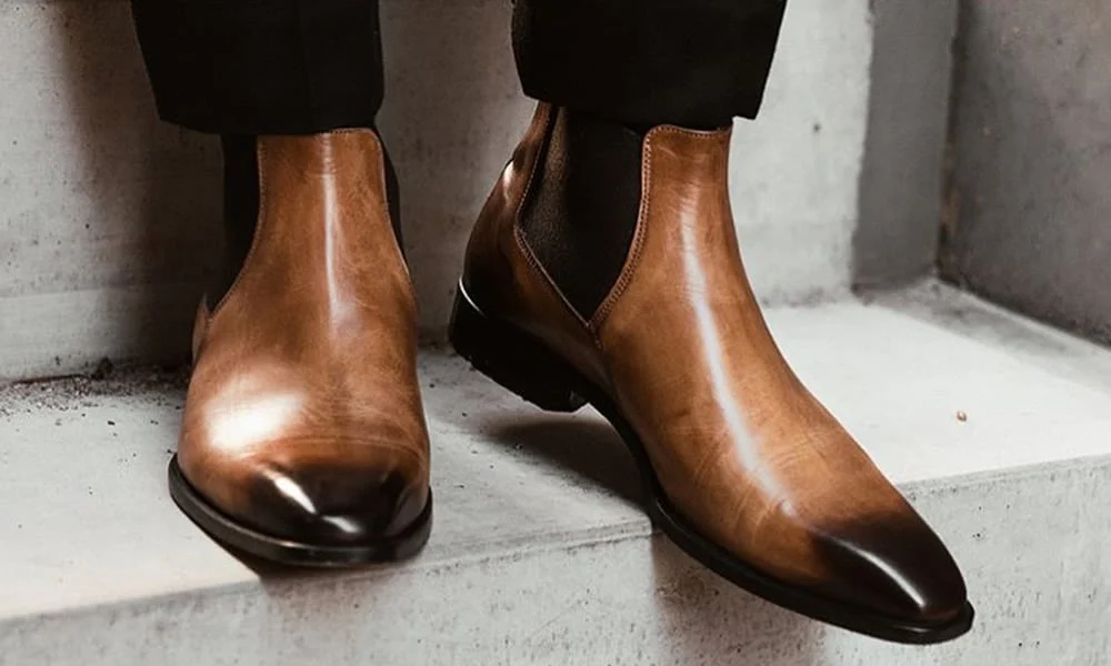 Polished tan leather Chelsea boots with dark elastic panels and tapered toes stand on a concrete step beneath black trousers, conveying sleek, refined style.