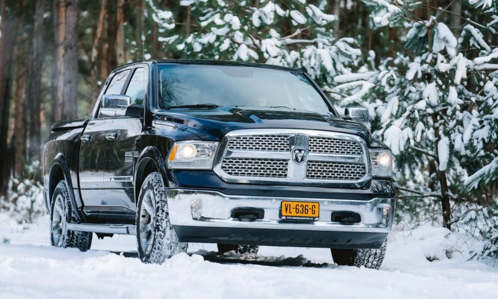 A black pickup truck with a chrome grille and yellow license plate drives through a snowy forest, framed by snow-covered evergreens; rugged and wintry.