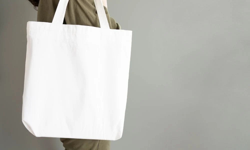 A person in olive clothing holds a large, plain white canvas tote bag against a soft gray background, creating a clean, minimalist product image.