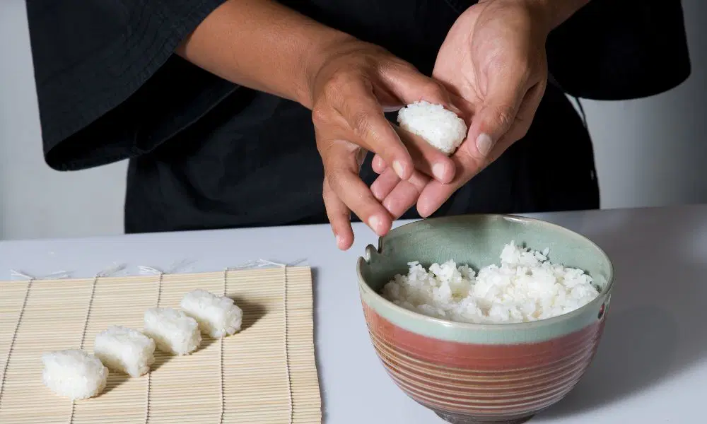 Hands shape a small ball of white rice over a ceramic bowl, with more rice balls arranged on a bamboo mat nearby; the scene feels calm and focused.