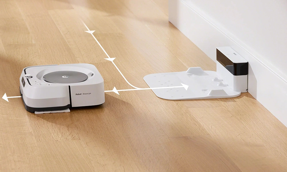 A white robot vacuum sits on a wood floor beside its charging dock, with arrows showing its route toward and away from the base.