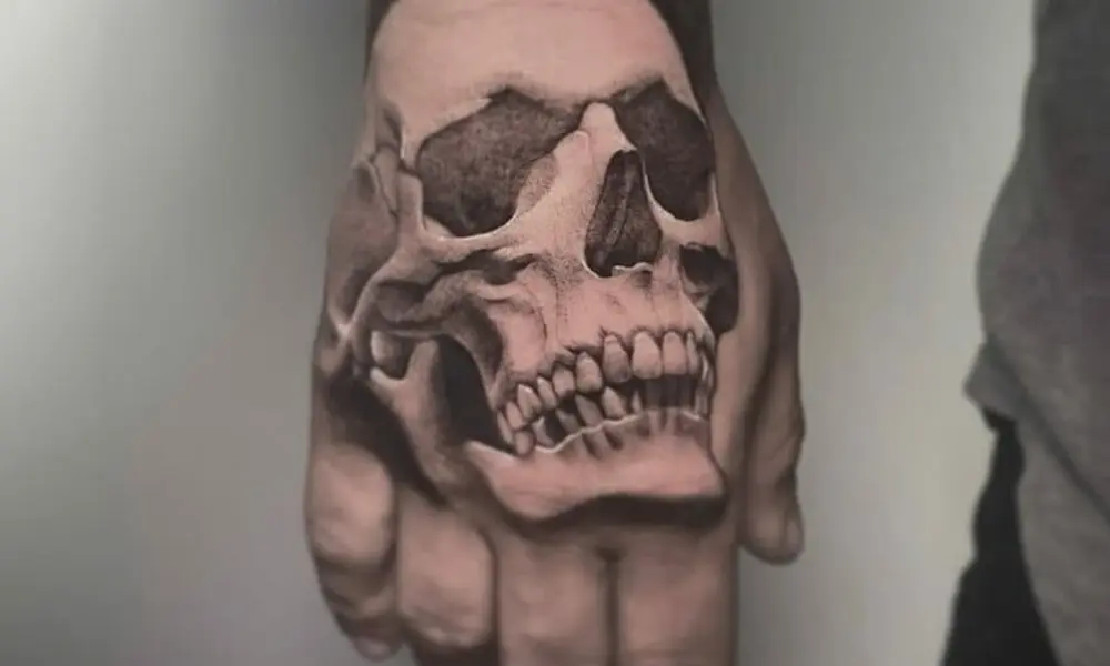 A hand tattoo of a grinning human skull spans the back of a clenched fist, creating a bold, dark, menacing effect against a plain wall.