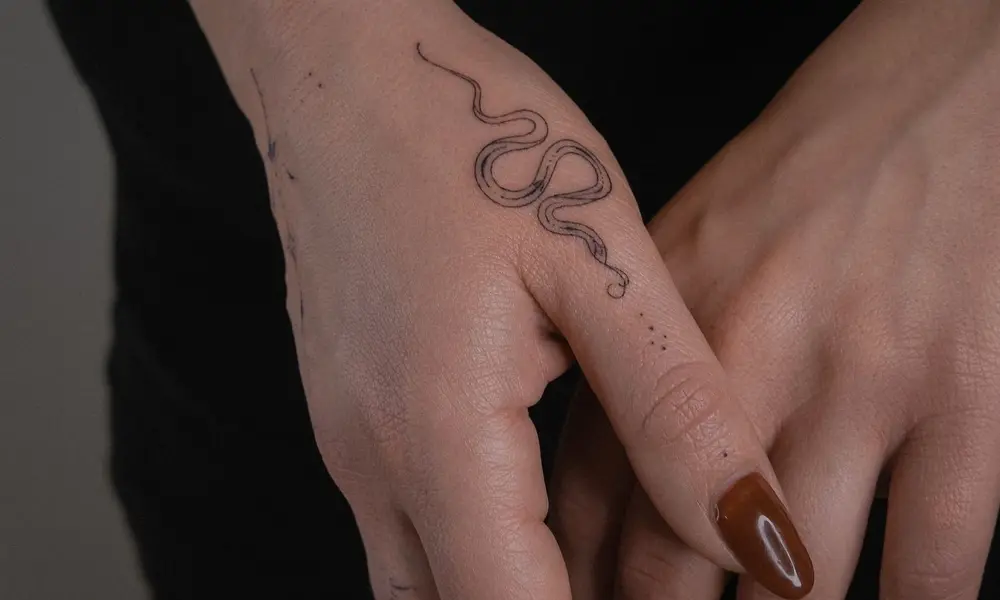 Close-up of two hands gently touching, one bearing a delicate black snake tattoo and glossy brown nail polish against a dark background.