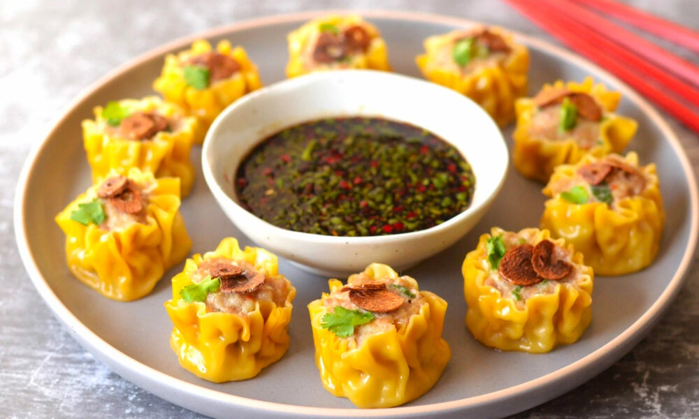 A plate of golden steamed shumai dumplings, topped with herbs and crisp bits, circles a bowl of dark chili dipping sauce; inviting food photography.