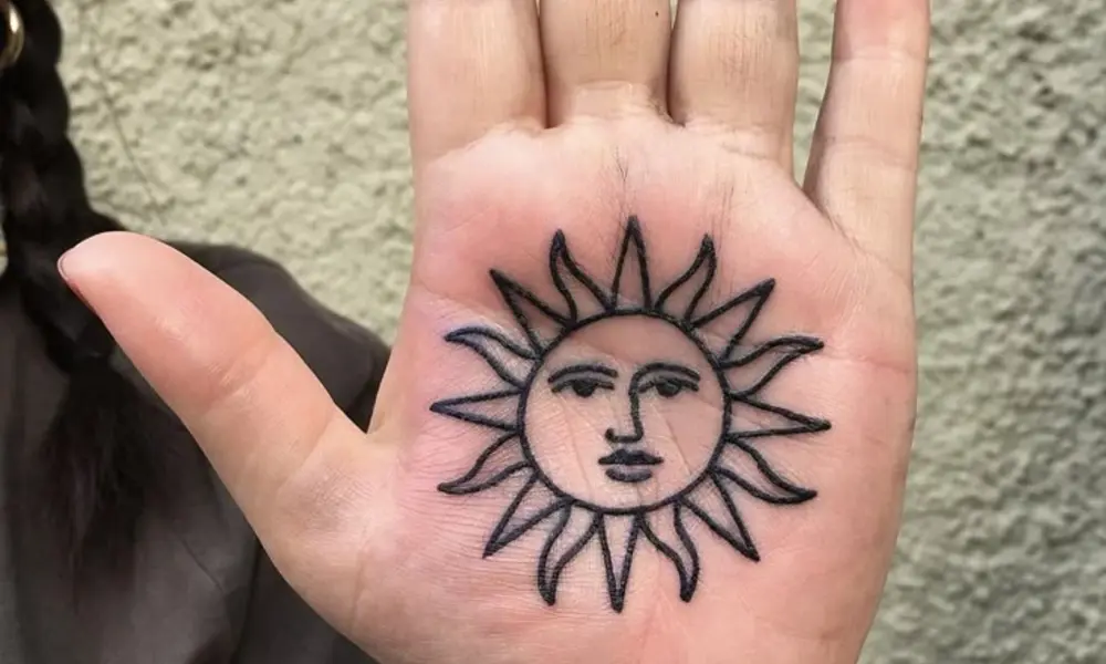 An open palm displays a black temporary tattoo of a sun with a calm human face and pointed rays, set against a softly textured outdoor background.