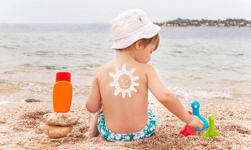 A young child in a white sunhat sits on the beach, a smiling sun painted on their back, with lotion and colorful sand toys nearby.