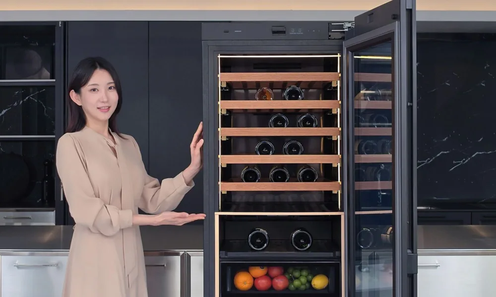 A smiling woman presents a built-in wine fridge filled with bottles and fruit in a sleek, dark modern kitchen, creating a warm, welcoming mood.