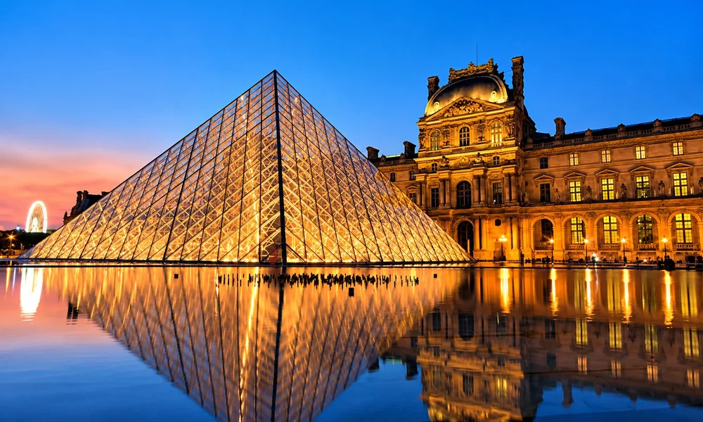 Glass pyramid and palace wings of the Louvre lit up at dusk, reflected in the water