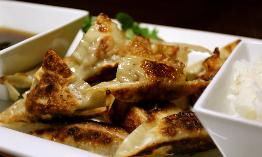 Golden-brown pan-fried dumplings with crisp, blistered wrappers fill a white platter, served with dipping sauce and rice for a warm, appetizing meal.